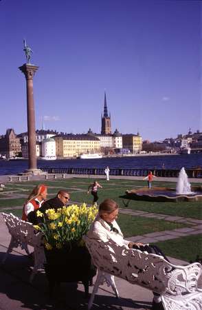 photo of Sweden, Stockholm, people sitting on white benches in the garden of the Stockholm City Hall (stadshuset) enjoying the view of Gamla Stan (Old Town) and the Swedish flag